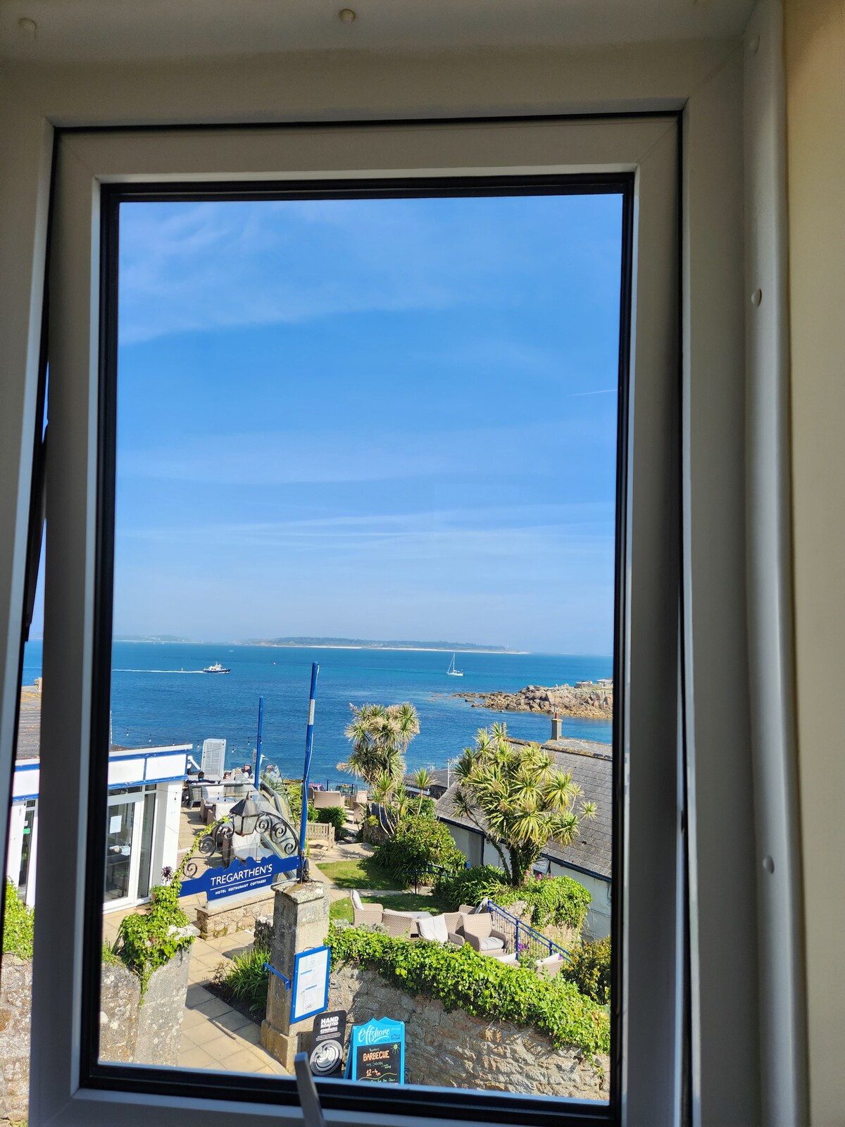 A large window frames a stunning sea view, revealing the vibrant blue water and a sailboat in the distance. Lush greenery and coastal elements are visible in the foreground, creating a serene atmosphere that captures the beauty of the surrounding landscape.