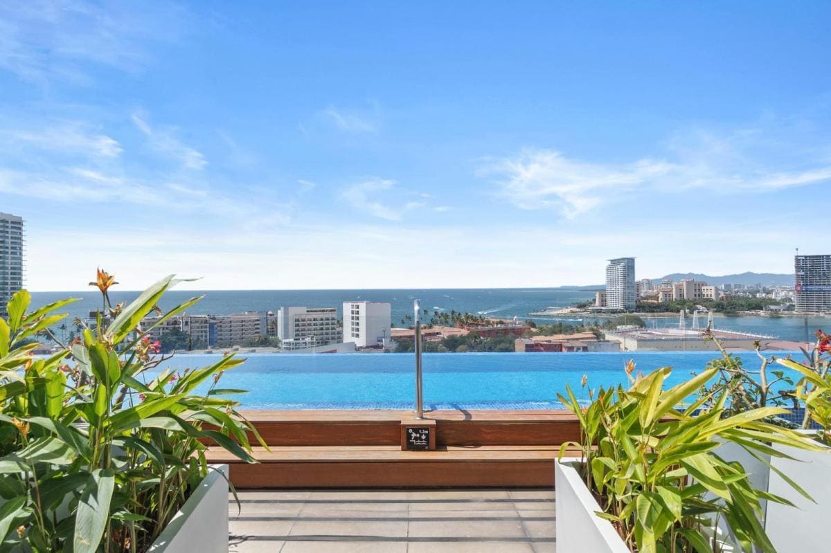 A rooftop pool offers expansive ocean views, framed by lush greenery. The horizon is lined with buildings and mountains, under a clear blue sky. The infinity edge of the pool blends seamlessly with the water, creating a serene and modern ambience.