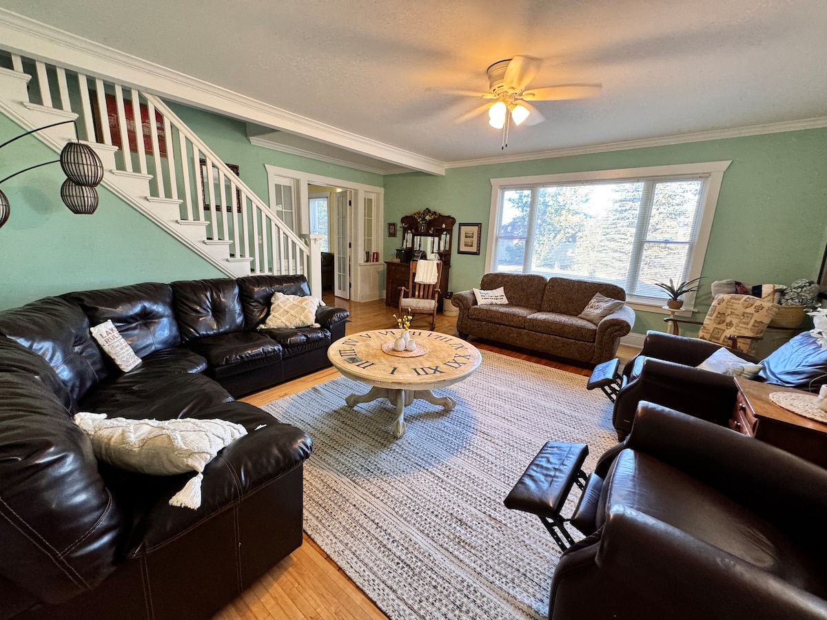 A spacious living area is furnished with a mix of leather sofas and comfortable chairs arranged around a round coffee table. Large windows provide ample natural light and a view of the surroundings. A textured area rug covers the hardwood floor.