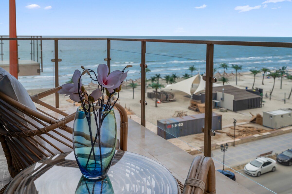 A view from the balcony captures the serene sea, with gentle waves lapping against the shore. A decorative vase holds delicate flowers on a glass table, complemented by stylish seating made of natural materials, allowing a relaxing atmosphere to enjoy the coastal scenery.