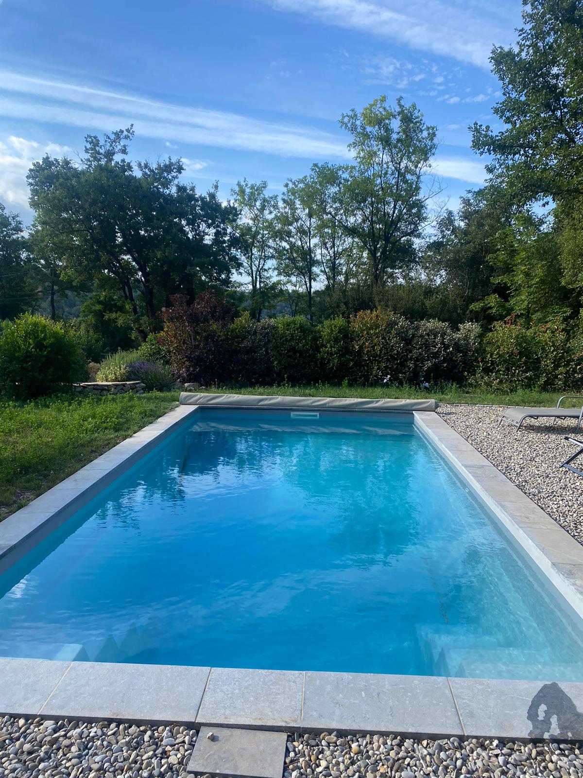 A private swimming pool is surrounded by a stone terrace and lush greenery. The clear water reflects the blue sky and trees, creating a serene outdoor environment. Sun loungers are positioned nearby, offering a comfortable space to relax.