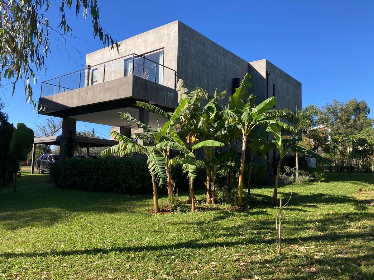 A modern two-story house features a sleek concrete exterior and large windows. Surrounded by greenery, including tropical plants, the structure is elevated, offering a clear view of the lush lawn. The bright blue sky enhances the vibrant atmosphere of the outdoor space.