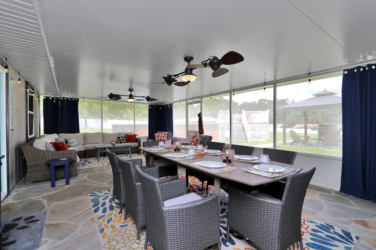 A spacious screened lanai features a long dining table surrounded by eight comfortable chairs. Relaxed seating is provided with a sectional sofa against the wall. Two ceiling fans are present, and large windows bring in natural light, creating an airy atmosphere. Outdoor views of the yard can be seen.