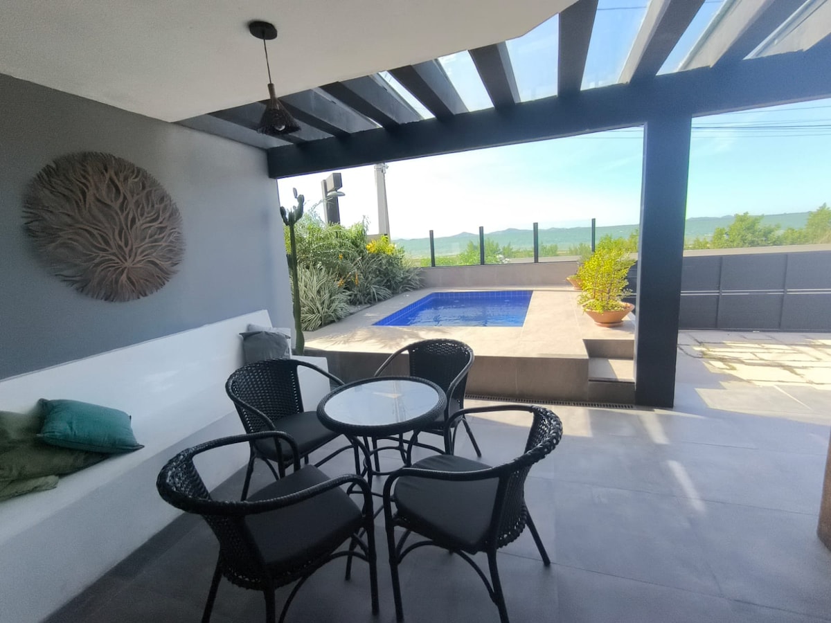 A covered outdoor seating area features a round table surrounded by four black wicker chairs. A modern plunge pool is visible nearby, with a scenic view of greenery and distant hills, framed by glass railings and a contemporary ceiling with exposed beams.
