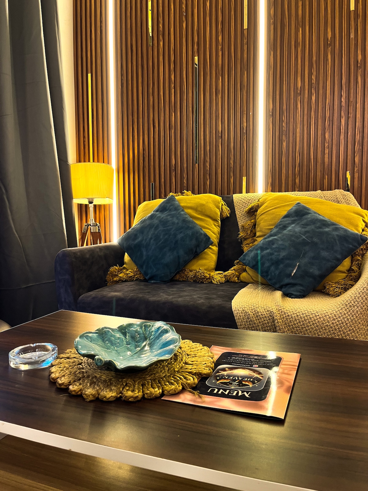 A comfortable seating area is presented with a dark sofa adorned with yellow and blue cushions. A decorative table is set with a blue dish and a glossy magazine, while a warm lamp casts soft light against wood-paneled walls.