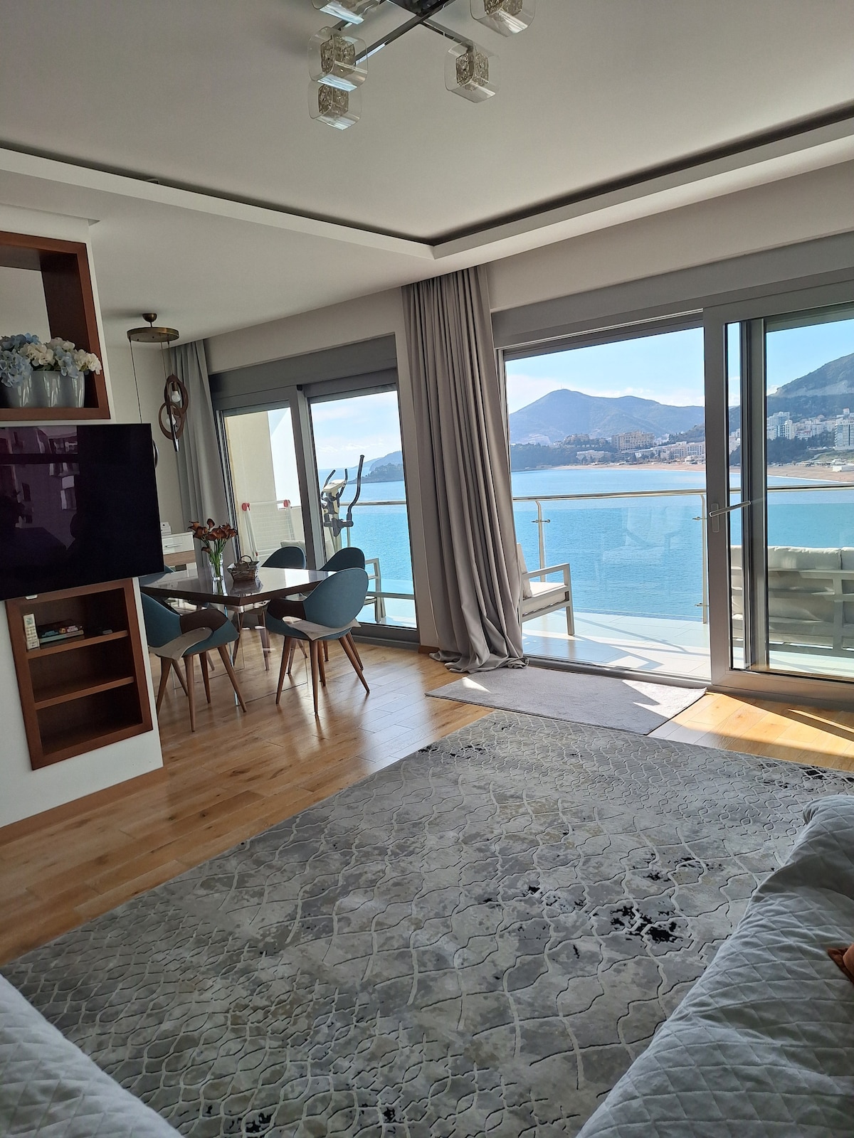 A bright living area is highlighted by large windows offering a view of the sea and mountains. Wooden flooring complements a light-colored rug, while a dining table with chairs is positioned near the windows. Natural light fills the room, enhancing the tranquil atmosphere.