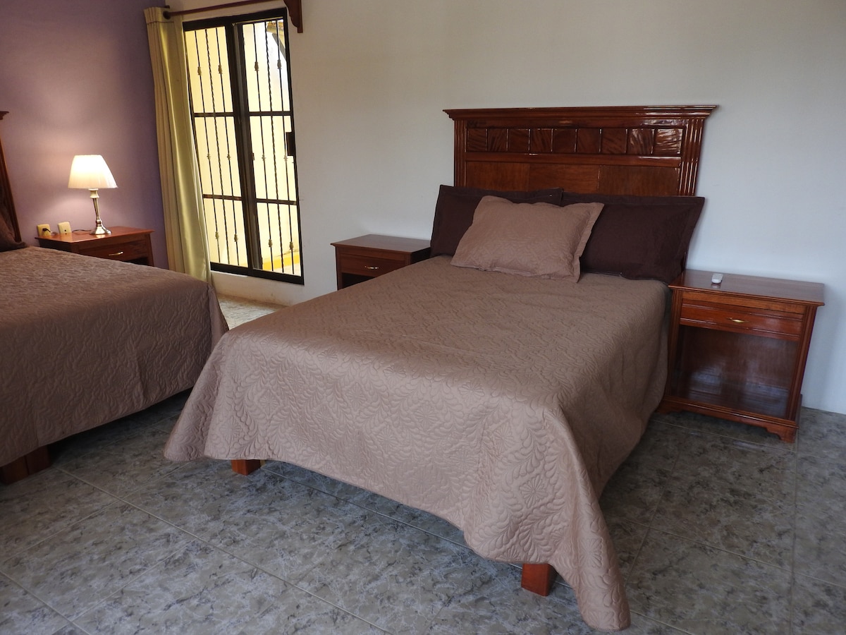 A spacious room is presented with two neatly made beds, each adorned with cozy beige bedspreads. Wooden nightstands sit adjacent to the beds, featuring lamps for soft lighting. A window with curtains allows natural light to enter, enhancing the welcoming ambiance.