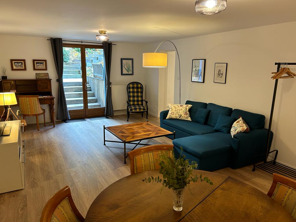 A spacious living area features a comfortable blue sectional sofa and a wooden coffee table. Natural light filters through large doors leading to an outdoor space. A round dining table is set for six, complemented by vintage wooden chairs. A warm glow comes from the pendant light.