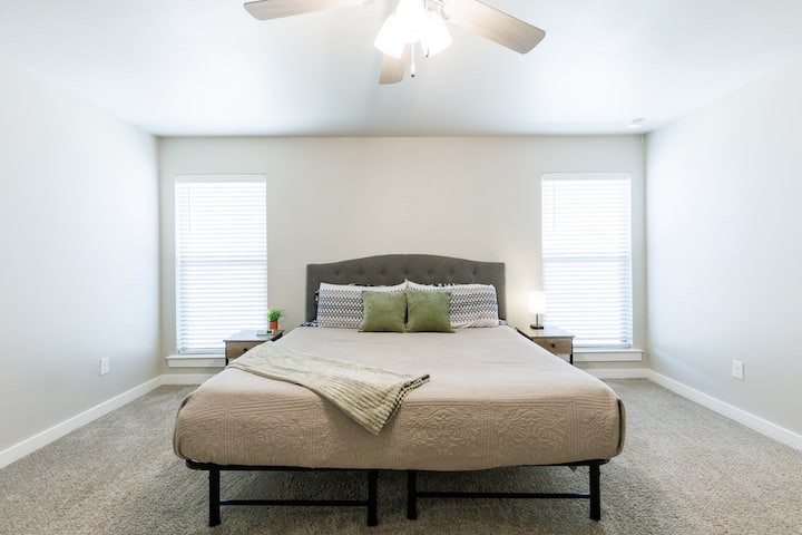 Táto spálňa má manželskú posteľ s neutrálnou prikrývkou a zelenými vankúšmi v spojení s rovnakými nočnými stolíkmi, oknami so žalúziami a stropným ventilátorom, ktoré sú umiestnené na podlahe pokrytej kobercom pre pokojnú a príjemnú atmosféru.