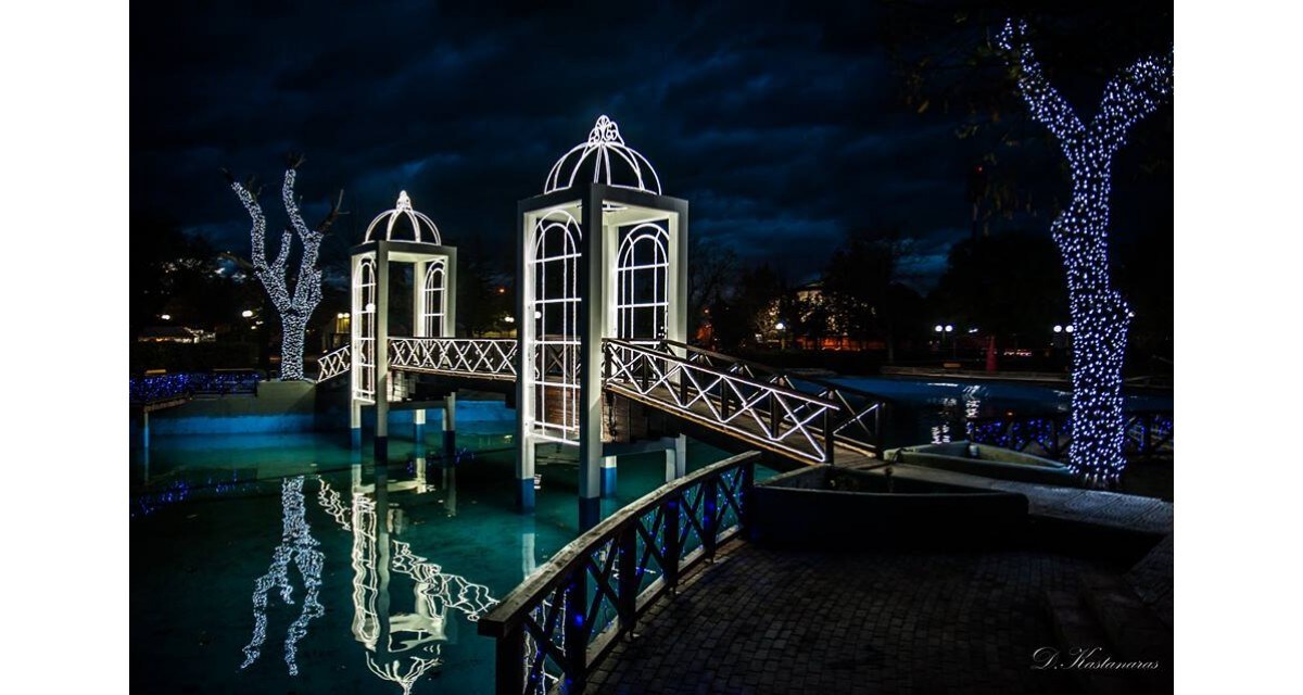 A serene view is captured at night, featuring a beautifully illuminated wooden bridge leading to two distinct structures. Surrounding trees are adorned with twinkling lights, reflecting softly on the calm water below, creating an inviting scene.