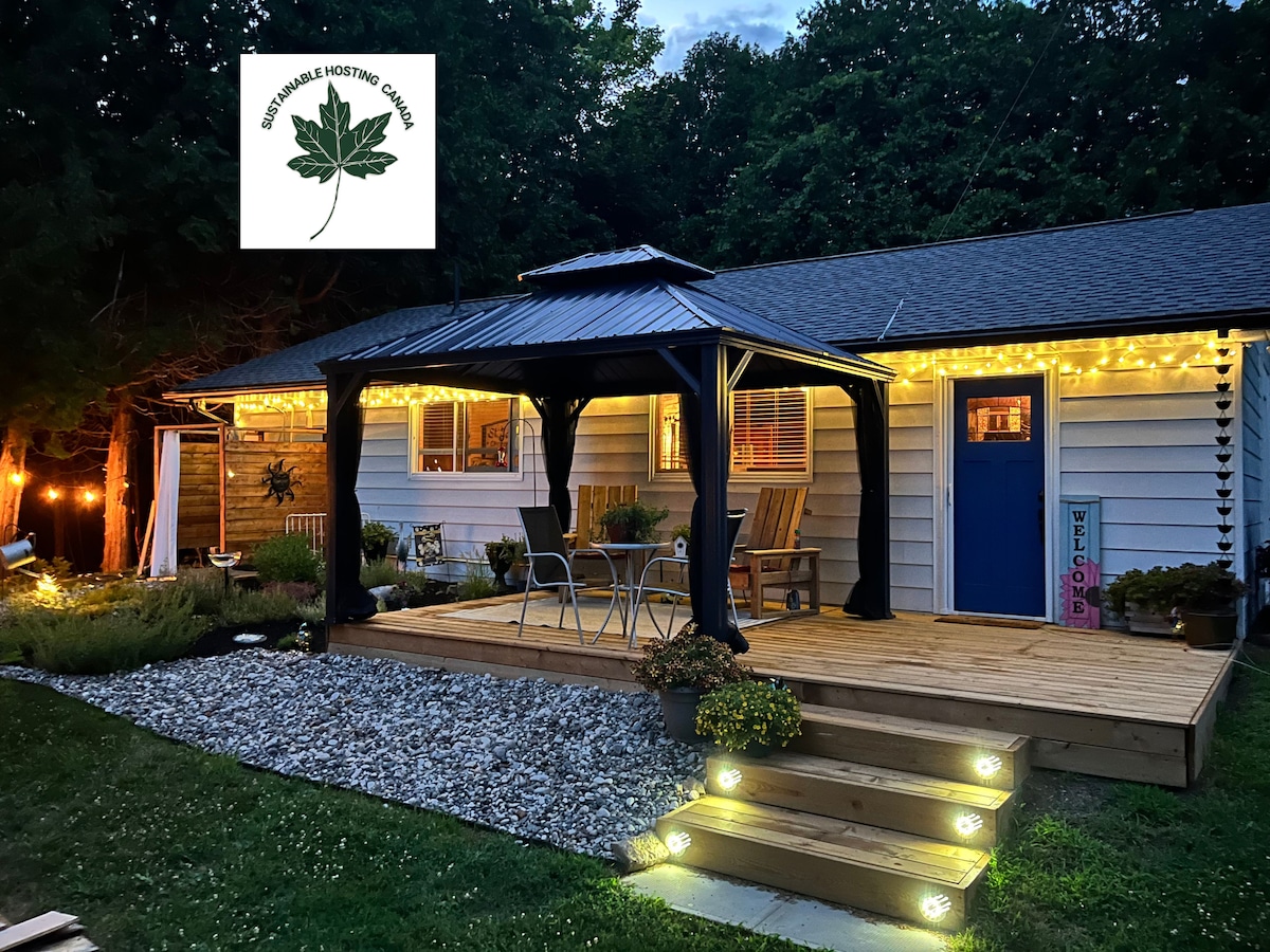 An inviting exterior view of the property features a wooden deck adorned with soft lighting. A covered seating area includes a small table and chairs, enhancing the outdoor space. Lush greenery surrounds the home, creating a tranquil setting for relaxation.