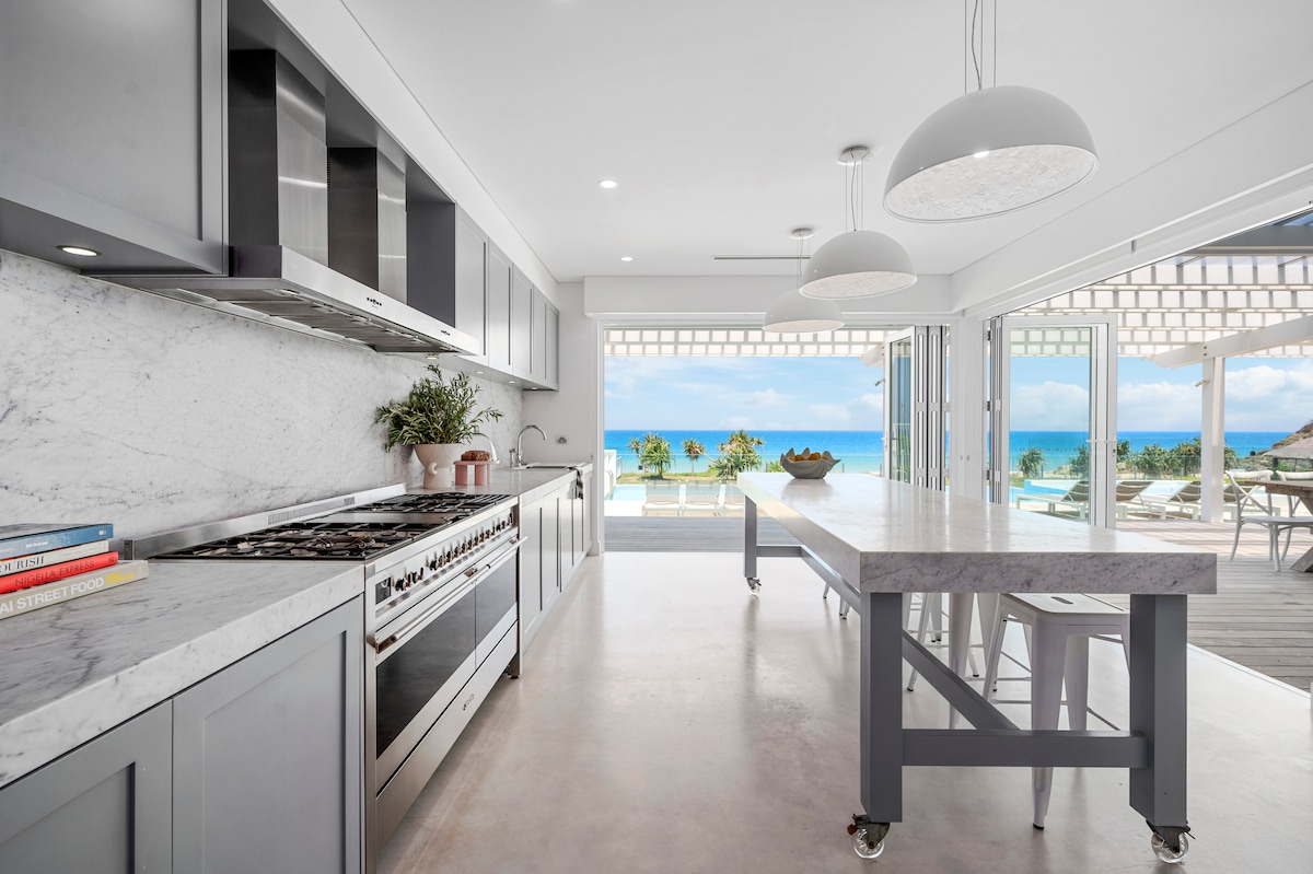 A modern kitchen features sleek gray cabinetry and a marble countertop. Stainless steel appliances are present, and a spacious island with seating is included. Large sliding glass doors provide a view of the outdoor area and ocean beyond, allowing natural light to fill the space.