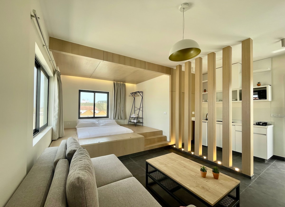 A modern studio space features a sleeping area with a spacious bed and a stylish sofa positioned across from it. Natural light enters through a large window, highlighting the warm wood accents and minimalist design. A functional kitchenette is visible in the background.