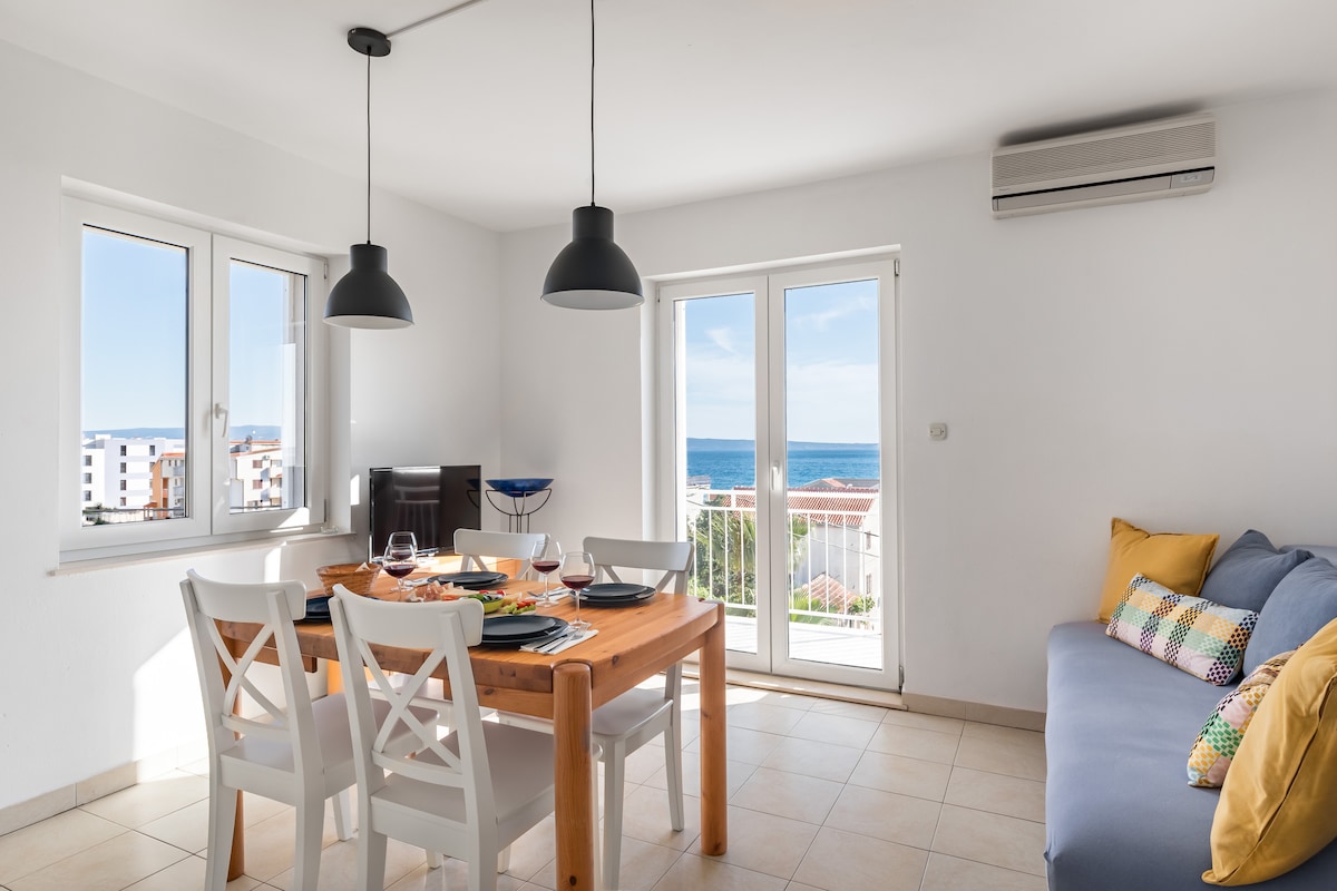 A dining area with a wooden table is accompanied by four white chairs. Two pendant lamps hang above the table. A blue sofa with colorful cushions is located nearby. Large windows provide natural light and offer a view of the sea and surrounding area.