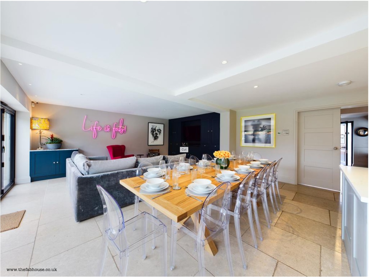 A spacious dining area featuring a large wooden table set for guests, surrounded by clear chairs. In the background, a cozy seating area is highlighted by soft furnishings and decorative elements. A neon sign adds a modern touch, while natural light streams through the windows.