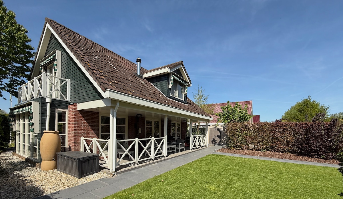 A spacious exterior view of the cottage features a well-kept yard with a green lawn and neatly arranged plants. The two-story structure displays a charming combination of wood and brick, complemented by a covered porch and a hot tub conveniently positioned nearby.