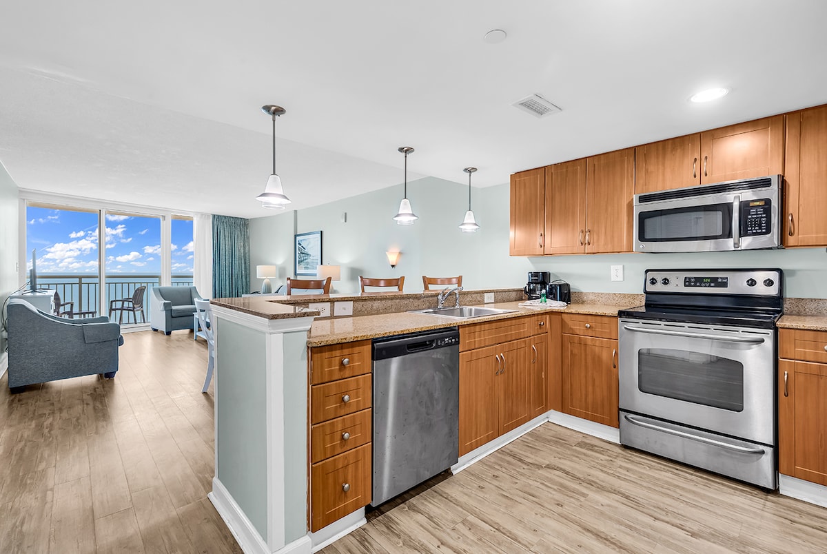 A modern kitchen is equipped with stainless steel appliances, including a stove and dishwasher, complemented by wooden cabinetry. The dining area features a table suitable for multiple guests. Large windows overlook the ocean, allowing ample natural light to fill the space.
