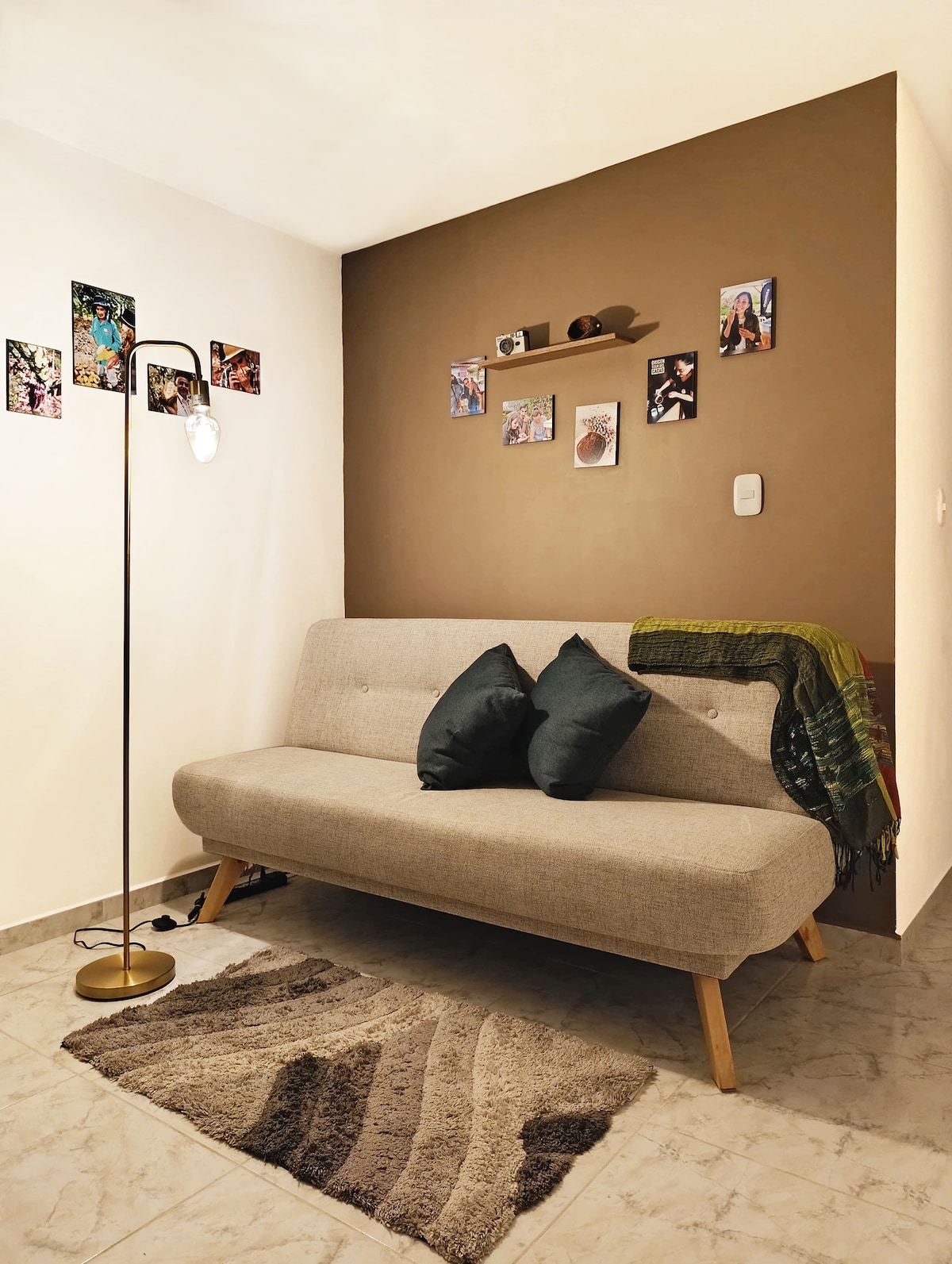 A cozy seating area features a light-colored sofa adorned with two dark pillows and a patterned throw. The wall behind is a rich brown, decorated with framed photos and a small shelf, while a slim standing lamp provides soft illumination.