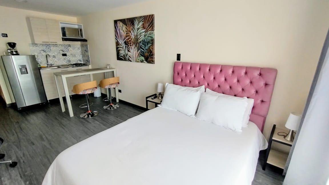 A spacious room features a double bed with white linens and a stylish pink upholstered headboard. A kitchenette area with bar stools and modern appliances is visible in the background. Decorative wall art adds a touch of color, while soft lighting creates a welcoming ambiance.