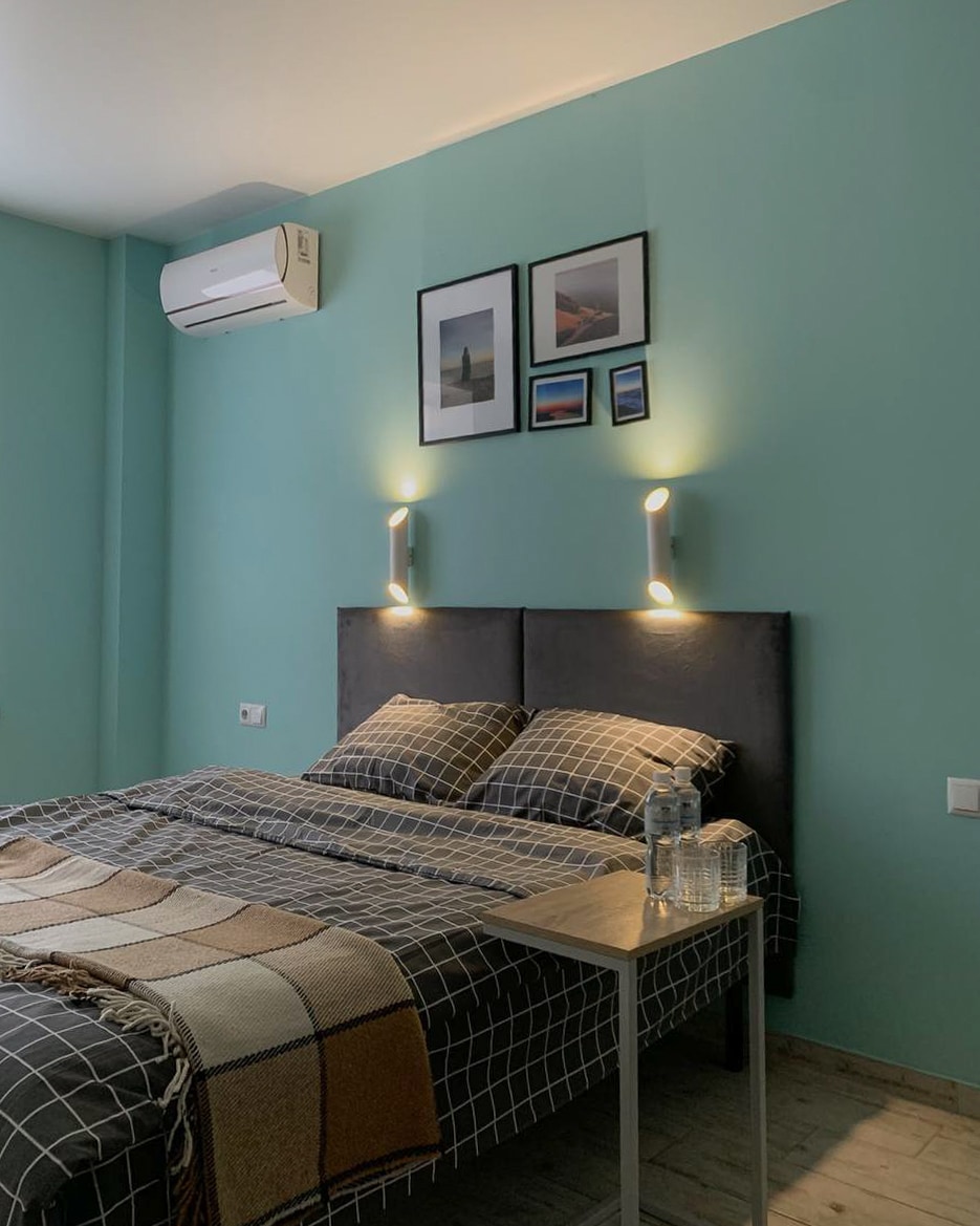 A sleeping area features a dark headboard with two wall-mounted lights. Above the bed, several framed photos are displayed. A checkered blanket is laid across a spacious bed, and a small table holds two bottles of water.