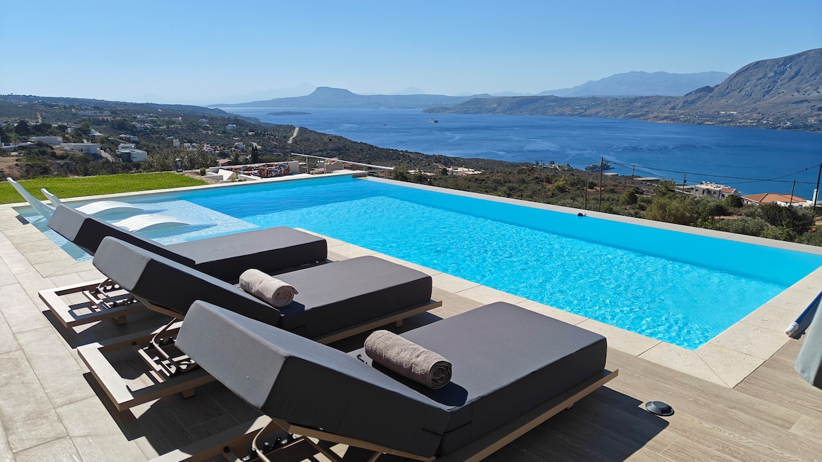 An infinity-edge swimming pool is surrounded by modern sun loungers with rolled towels placed on each. The scene overlooks scenic views of a calm sea, distant mountains, and a clear blue sky, creating a serene atmosphere for relaxation.