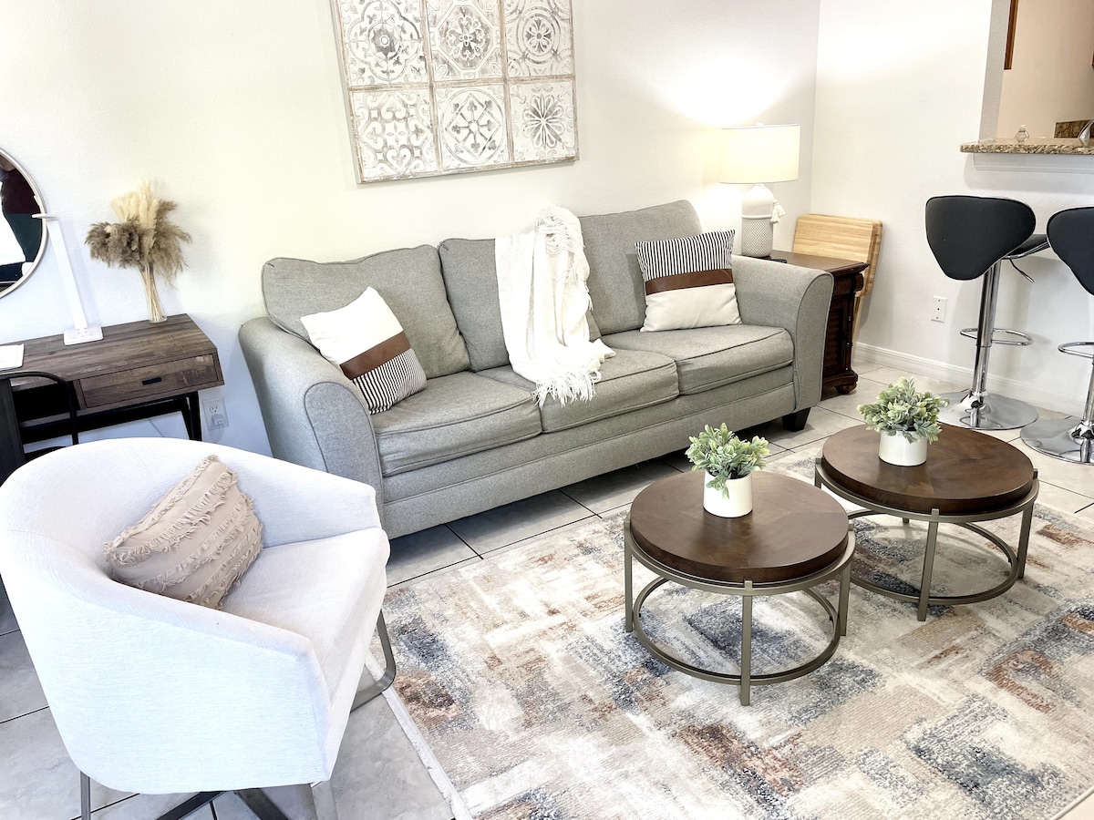 A cozy seating area features a grey sofa along with two round coffee tables adorned with potted plants. The white armchair complements the space, which is softly illuminated by a lamp. A neutral-toned rug adds warmth to the tile flooring.
