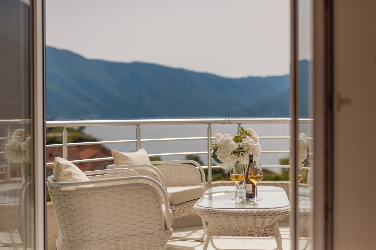 A cozy outdoor seating area features two white wicker chairs and a glass-topped table, set against a backdrop of mountains and a tranquil bay. Fresh flowers are placed on the table, while sunlight highlights the inviting space.