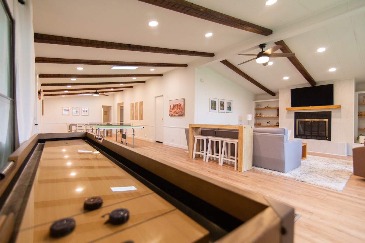 A spacious living area features an inviting layout with a shuffleboard table in the foreground. The room is illuminated by recessed lighting, and a large sectional sofa is positioned near a stone fireplace. A television is mounted above a wooden shelf, providing entertainment options.