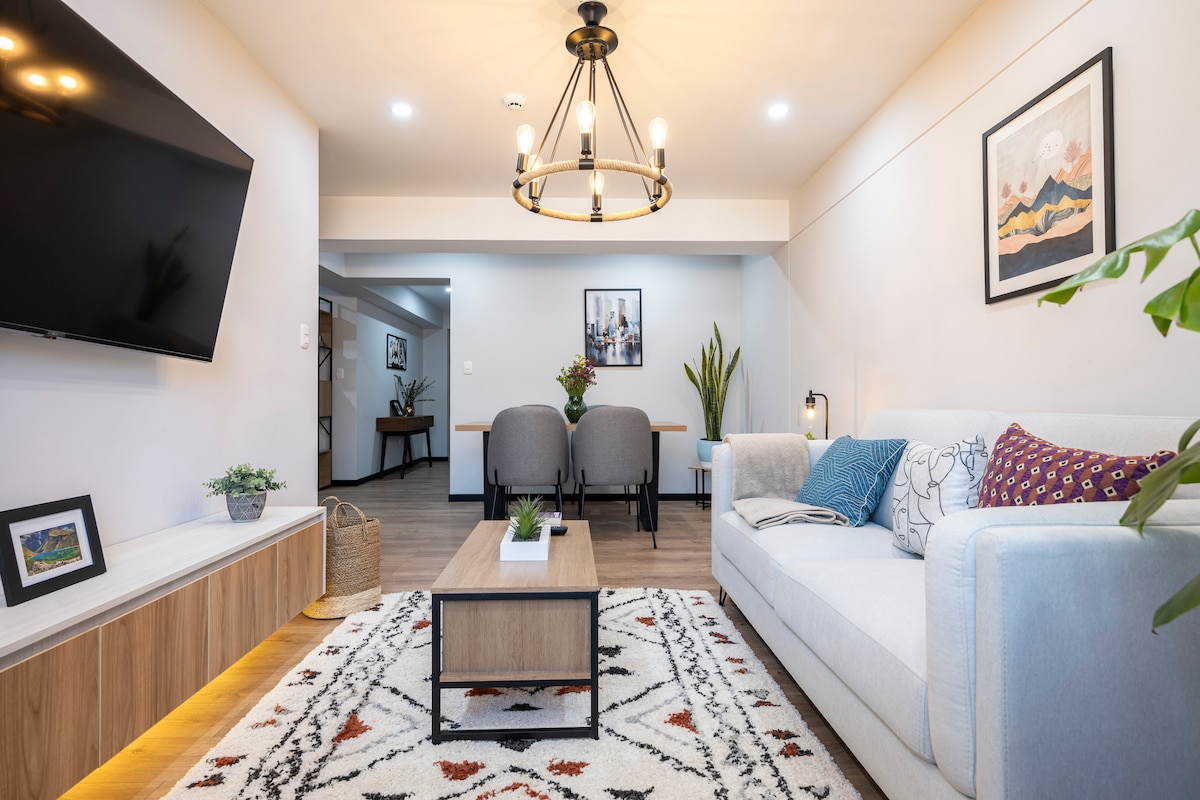 A modern living room is presented with a comfortable sofa and a large television mounted on the wall. A coffee table rests on an area rug, complemented by two stylish chairs nearby. Plants add a touch of greenery, enhancing the welcoming ambiance.
