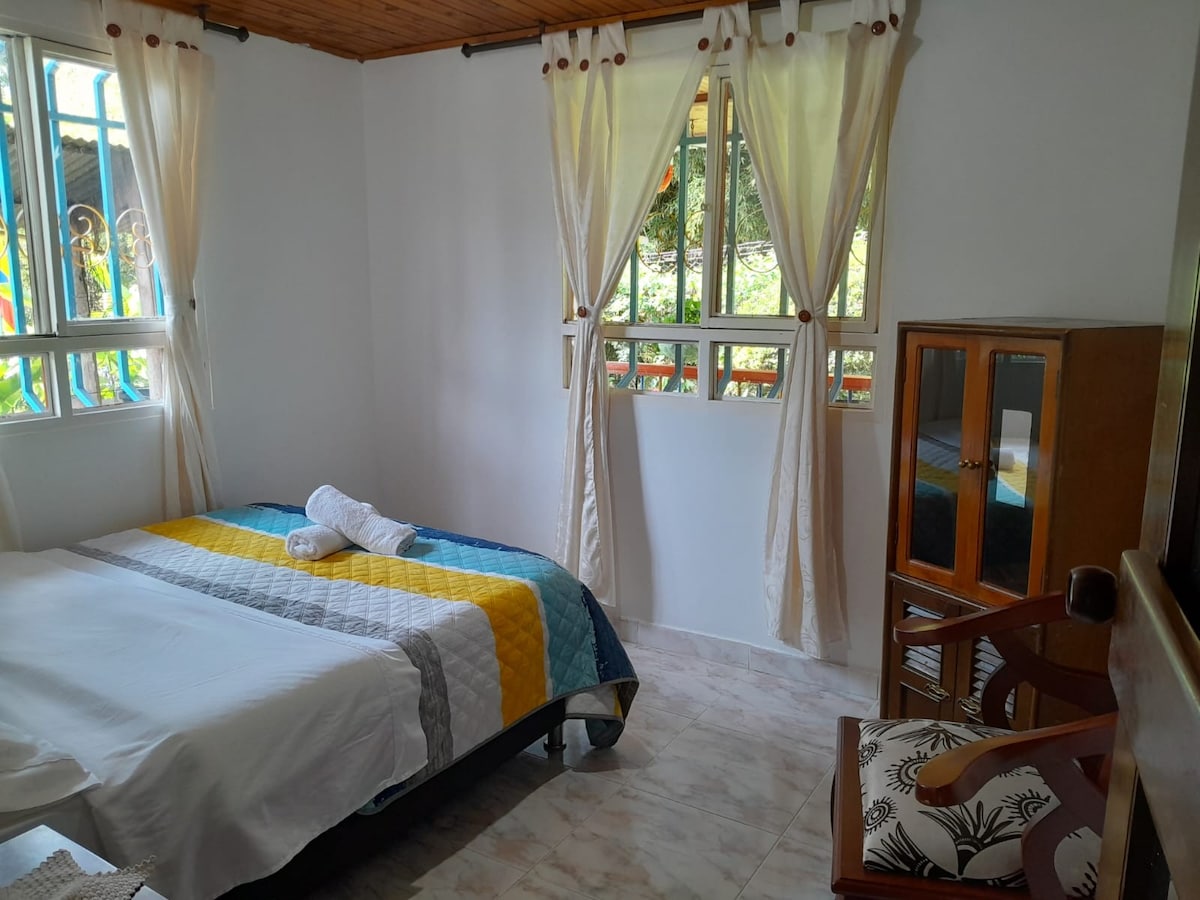 A bright sleeping area features a spacious bed with a colorful quilt, complemented by fresh towels. Natural light streams through multiple windows, illuminating the room. Nearby, a wooden cabinet and a chair add to the functional decor, creating a cozy environment.