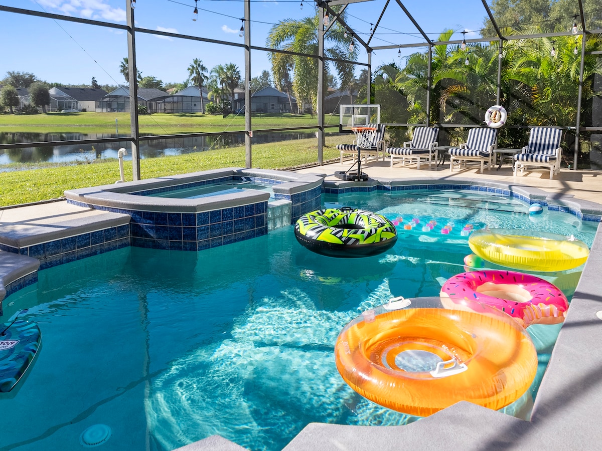 A private pool area is enclosed by a screened lanai, featuring a hot tub beside the pool. Colorful floating rings in various shapes rest on the water, alongside a backdrop of lush tropical plants and a peaceful pond view in the distance.