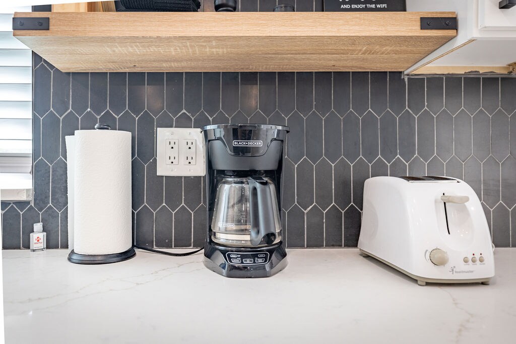 A modern kitchen countertop displays a coffee maker and a toaster, both in contrasting colors. A white paper towel holder is positioned nearby, with a small decorative item visible. A textured backsplash adds depth to the space, enhancing the overall design.