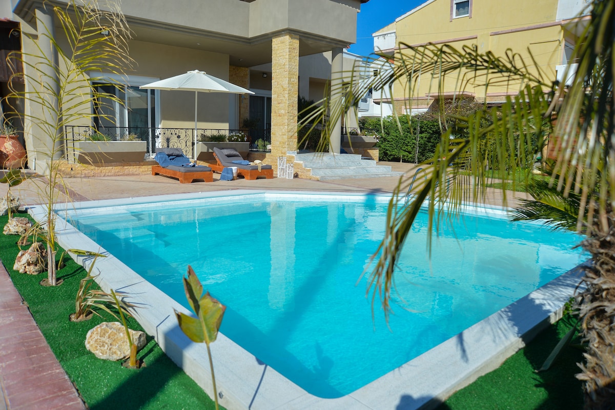 A serene outdoor area features a sparkling pool surrounded by well-maintained greenery. Comfortable lounge chairs are positioned nearby under a shaded umbrella. The spacious patio provides a welcoming space for relaxation, with the home's exterior visible in the background.