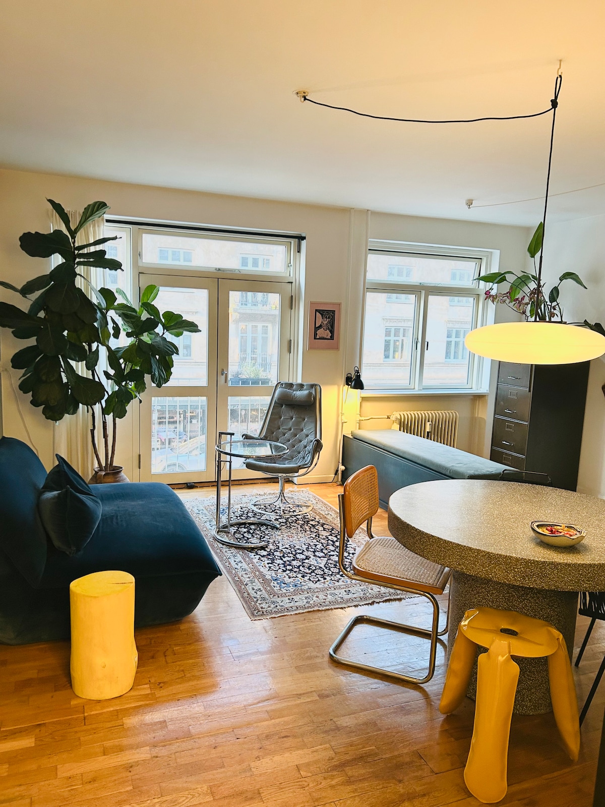 A living area is featured with a plush couch and a textured armchair, both positioned near large windows that allow ample natural light. A circular table sits near a vibrant area rug, with a decorative bowl atop it. Greenery adds a refreshing touch to the space.