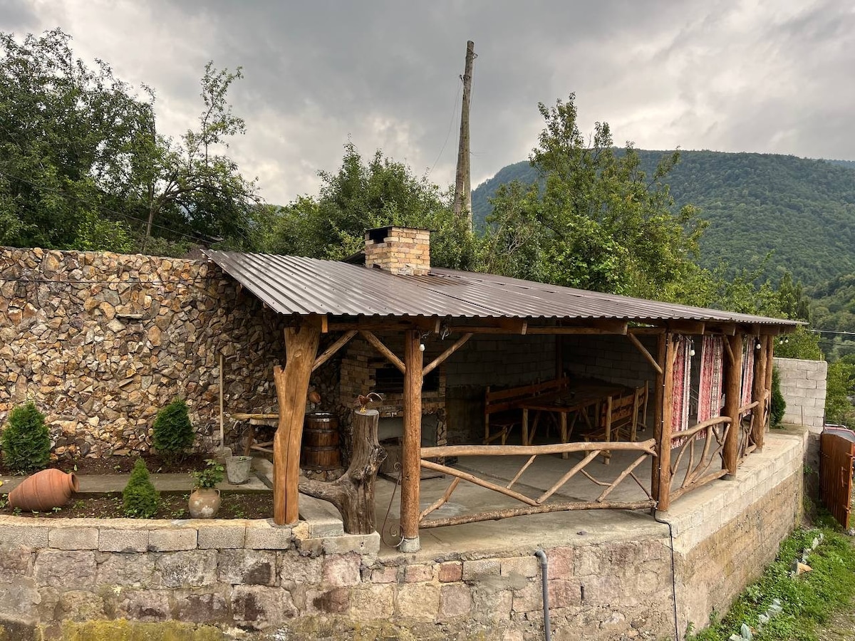 An outdoor seating area is featured under a rustic wooden roof, supported by natural wood beams. Stone walls provide a sturdy backdrop, while trees and shrubs surround the space, contributing to a serene environment. A large wooden table and benches are visible, inviting gatherings.