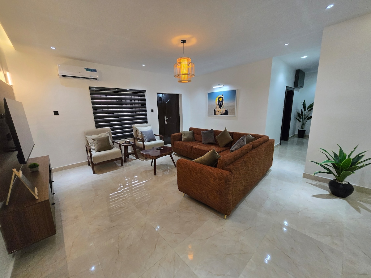 A spacious living area is highlighted by a large, comfortable couch and two upholstered chairs. A wooden coffee table occupies the center, while a wall-mounted TV is positioned across from the seating. Soft lighting enhances the modern decor, complemented by indoor plants and elegant wall art.