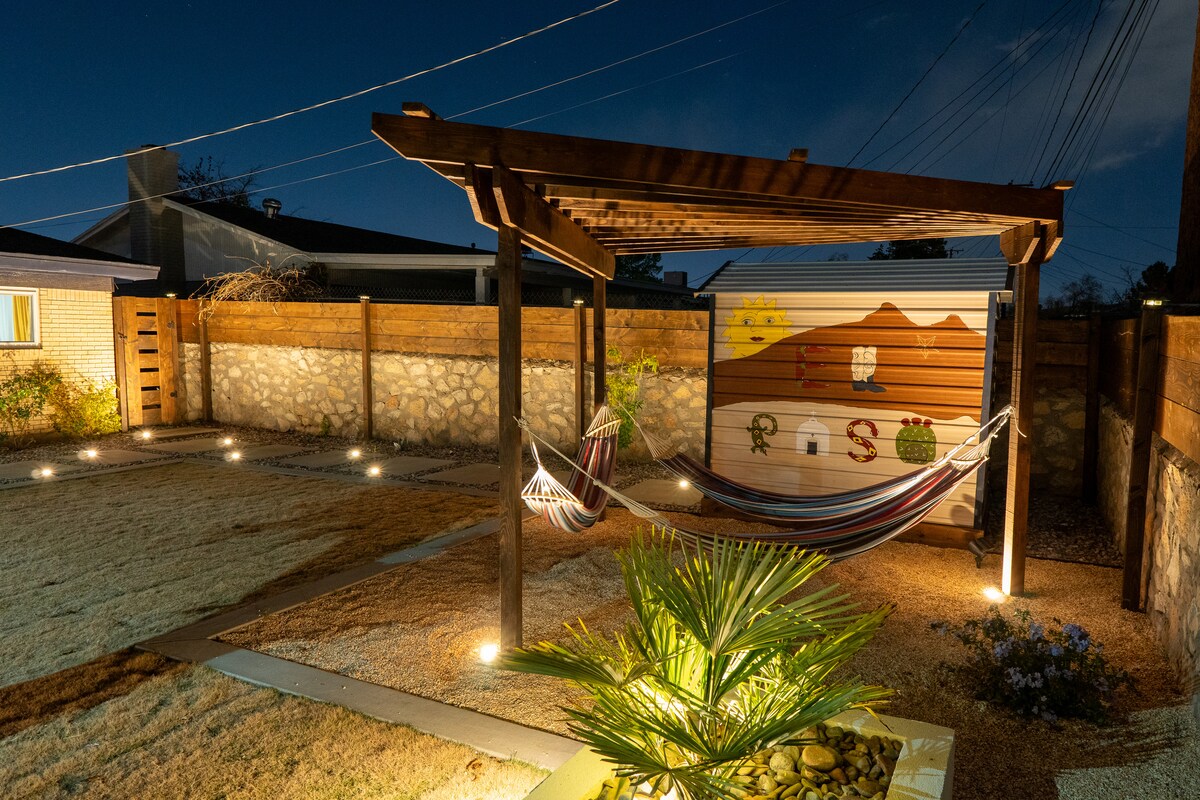 A backyard retreat features a wooden pergola supporting a colorful hammock beneath soft outdoor lighting. Surrounding plants accentuate the tranquil space, with stone pathways leading to a basketball court. The serene atmosphere is complemented by painted accents on the wall, enhancing the cultural theme.