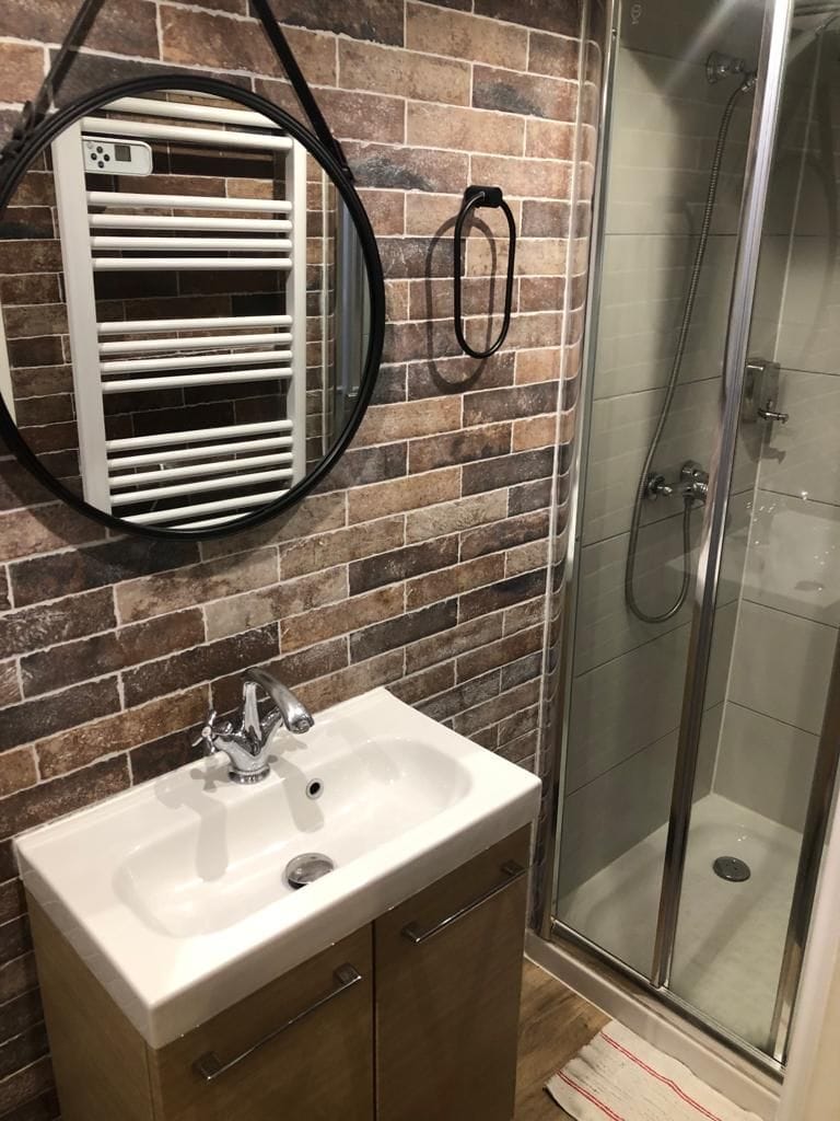 A modern bathroom features a sleek sink set within a wooden cabinet. A round mirror is mounted above the sink, and a towel heater is positioned on the wall. The shower area is enclosed with glass, surrounded by stylish tilework that complements the overall design.