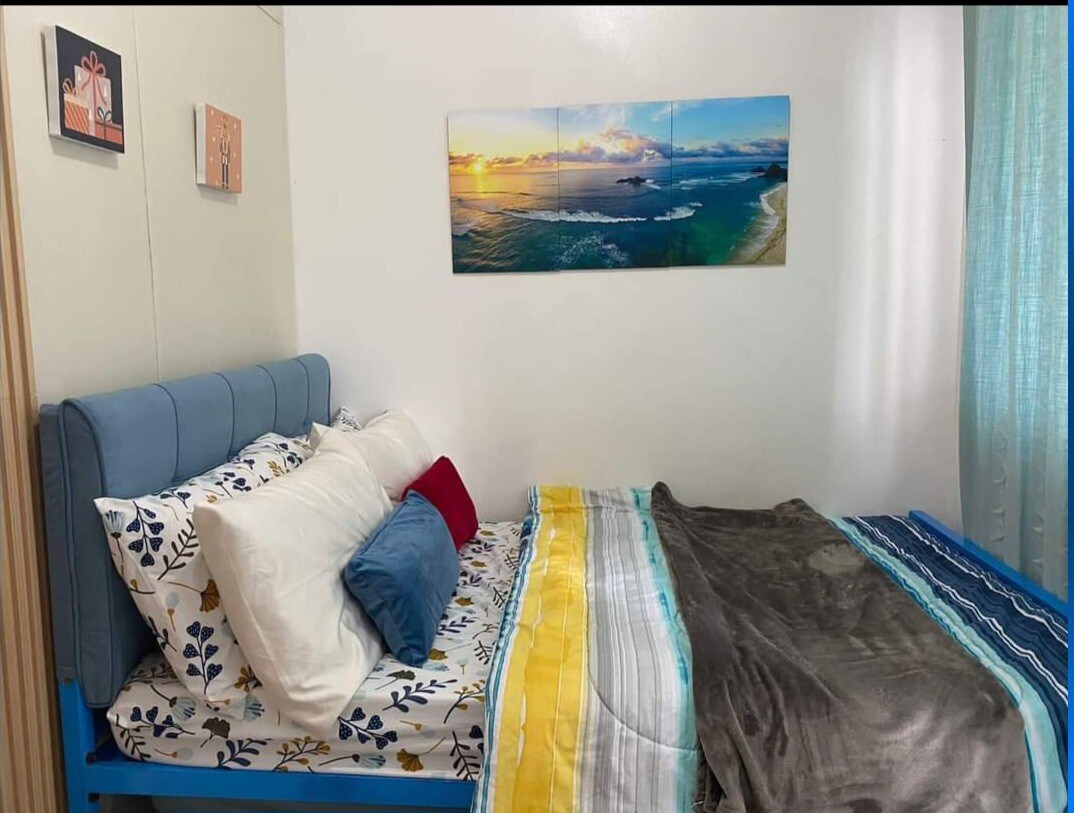 A cozy sleeping area features a blue-framed bed with layered bedding in various colors, including a grey throw blanket and decorative pillows. A beach-themed artwork hangs above the bed, complemented by soft natural light filtering through sheer curtains.