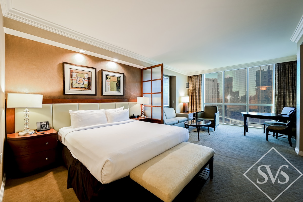 The spacious bedroom features a king-sized bed with crisp white linens and a beige upholstered bench at the foot. Natural light floods the room through large windows, showcasing views of the city. A divider separates the sleeping area from a cozy seating space with a loveseat and table.