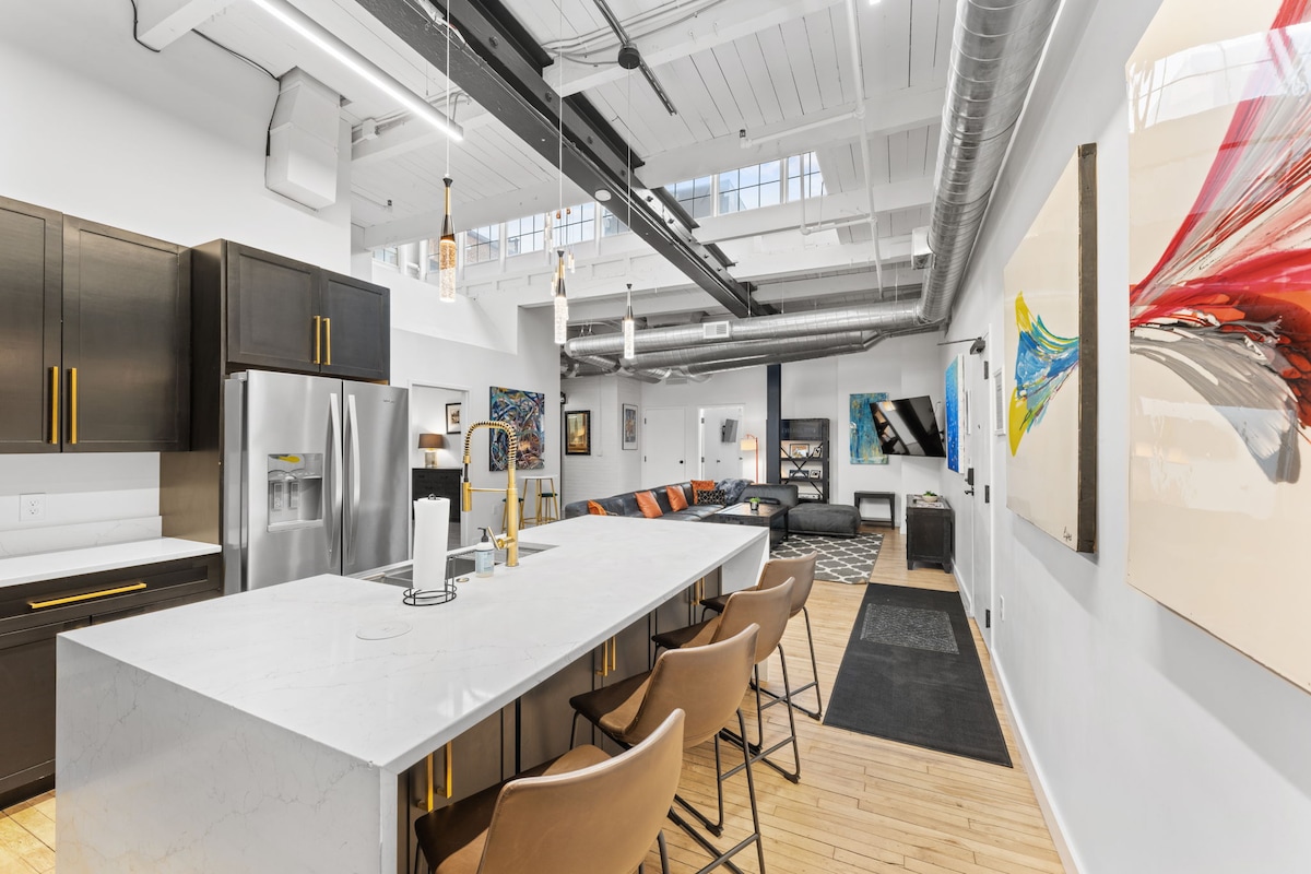 An open kitchen area is highlighted by a large marble island with seating for four. Dark cabinetry contrasts with light countertops. The ceiling is adorned with exposed beams and modern light fixtures, while art pieces and a comfortable living area are visible in the background.