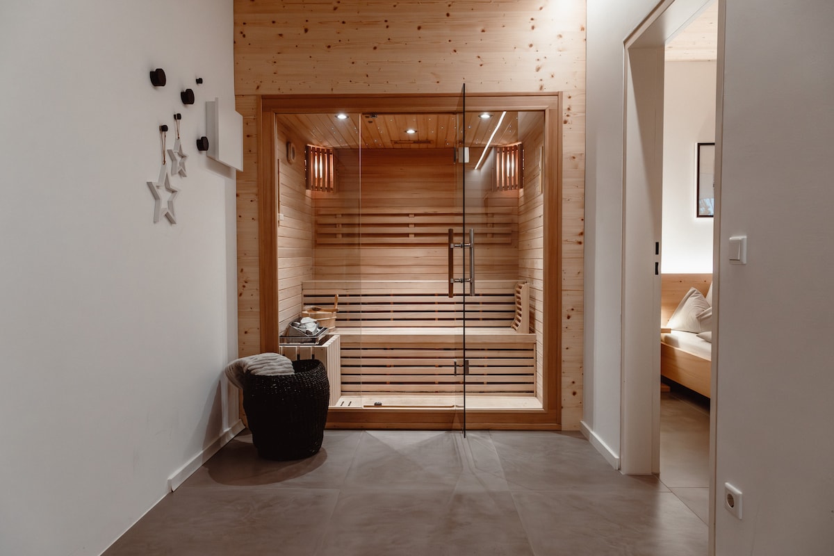A modern sauna area is framed by wooden walls and features a glass door. Subtle lighting highlights the wood slats, creating a tranquil atmosphere. A small seating area with a woven basket is visible, inviting relaxation after a day of outdoor activities.