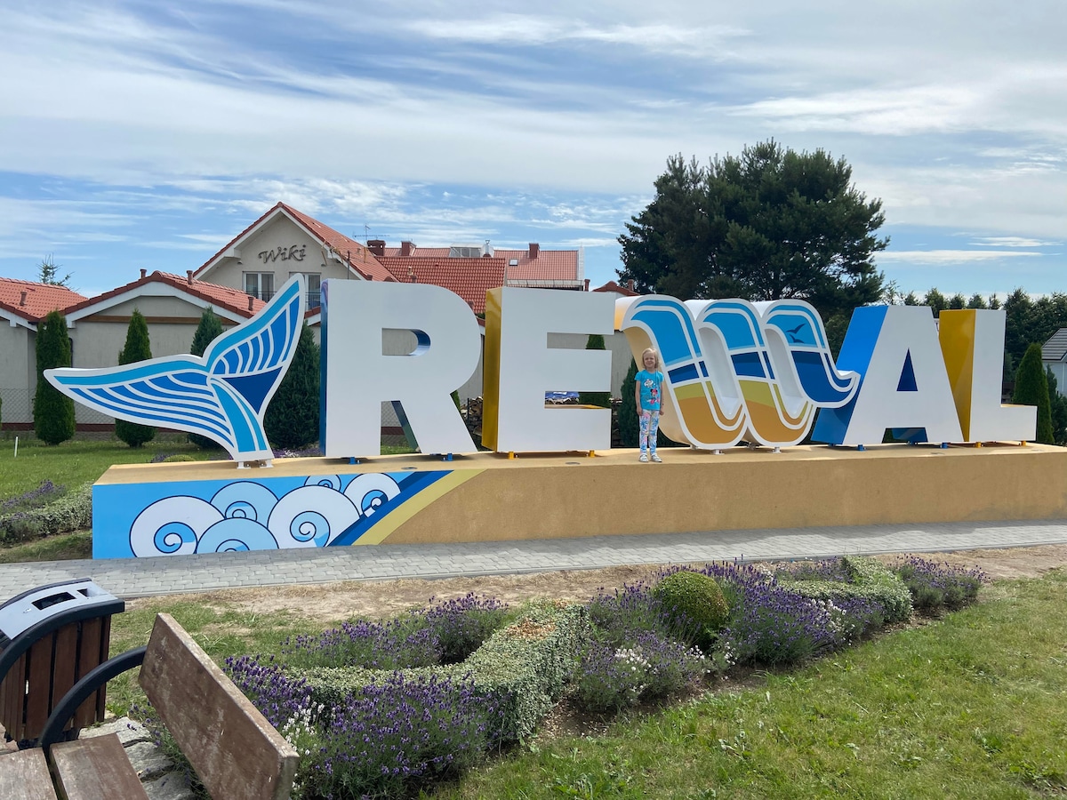 A large sign displaying the name 'Rewal' stands prominently, featuring vibrant colors and artistic designs. The structure is complemented by decorative waves and birds, set against a backdrop of greenery and buildings, inviting visitors to the area.