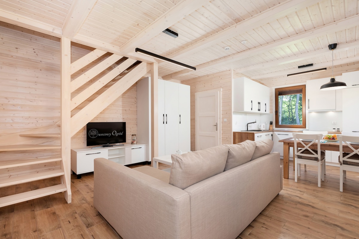 A spacious interior features wooden walls and ceiling, creating a warm environment. A comfortable sofa is positioned in front of a television, while a kitchen and dining area are visible in the background. Natural light illuminates the room through a nearby window.