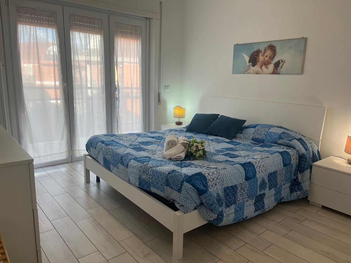 A well-lit bedroom features a spacious bed adorned with a blue-patterned quilt. Two bedside lamps provide soft lighting. Large glass doors lead to a balcony, dressed with sheer curtains that allow natural light to filter in.