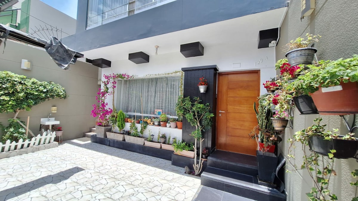 A welcoming entryway is framed by vibrant potted plants and climbing flowers. The door is made of wood, flanked by windows that allow natural light to fill the space. The floor features patterned tiles, enhancing the outdoor ambiance.