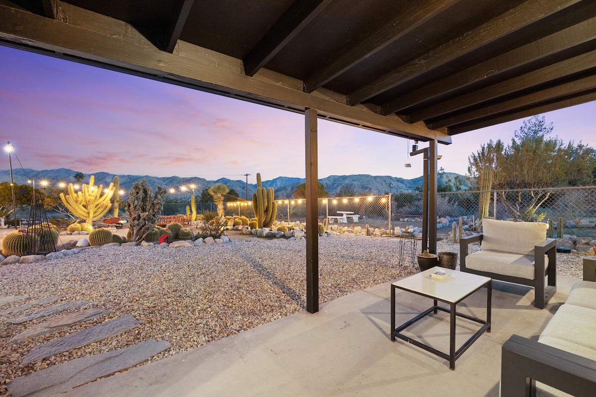 A covered outdoor seating area presents a modular couch and a low table, surrounded by decorative gravel and cacti. An expansive view of the desert landscape and mountains is visible, complemented by soft lighting that adds to the serene ambiance as dusk approaches.