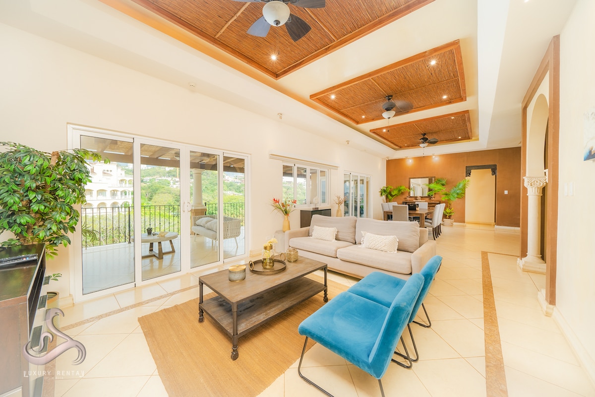 A spacious living area features a large sectional sofa, complemented by a blue accent chair. Multiple sliding glass doors provide natural light and open to a balcony with scenic views. Elegant ceiling fans circulate air in the welcoming space, enhancing comfort.