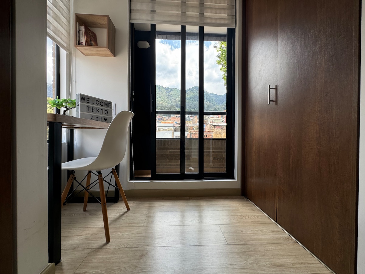 A work area is seen featuring a white chair positioned at a wooden desk. Large floor-to-ceiling windows allow natural light to illuminate the space, offering a glimpse of the mountainous backdrop outside. A decorative welcome sign is displayed on the desk.