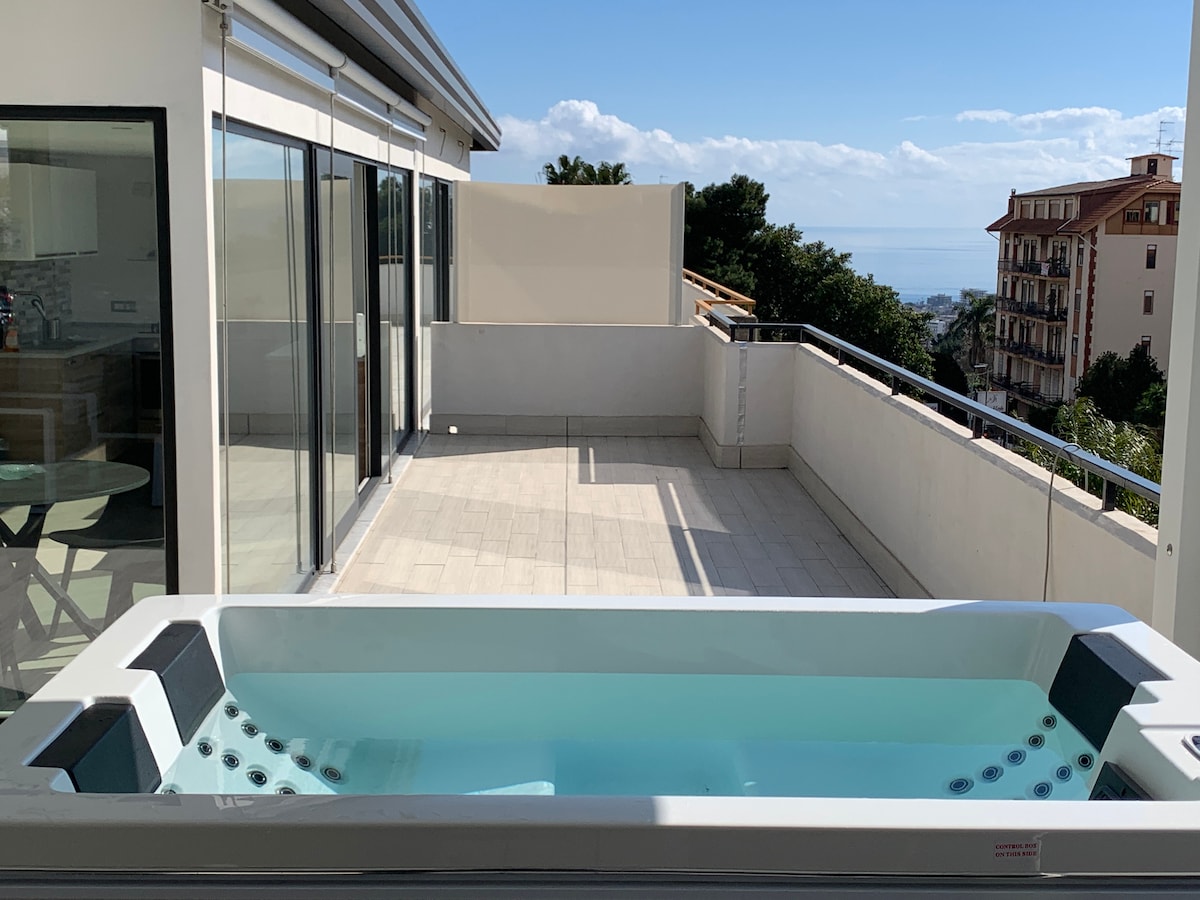A spacious terrace featuring a private jacuzzi is shown, overlooking a cityscape. Large glass doors provide abundant natural light, and views of the distant sea can be seen through the open railing. The tiled floor adds to the modern ambiance.